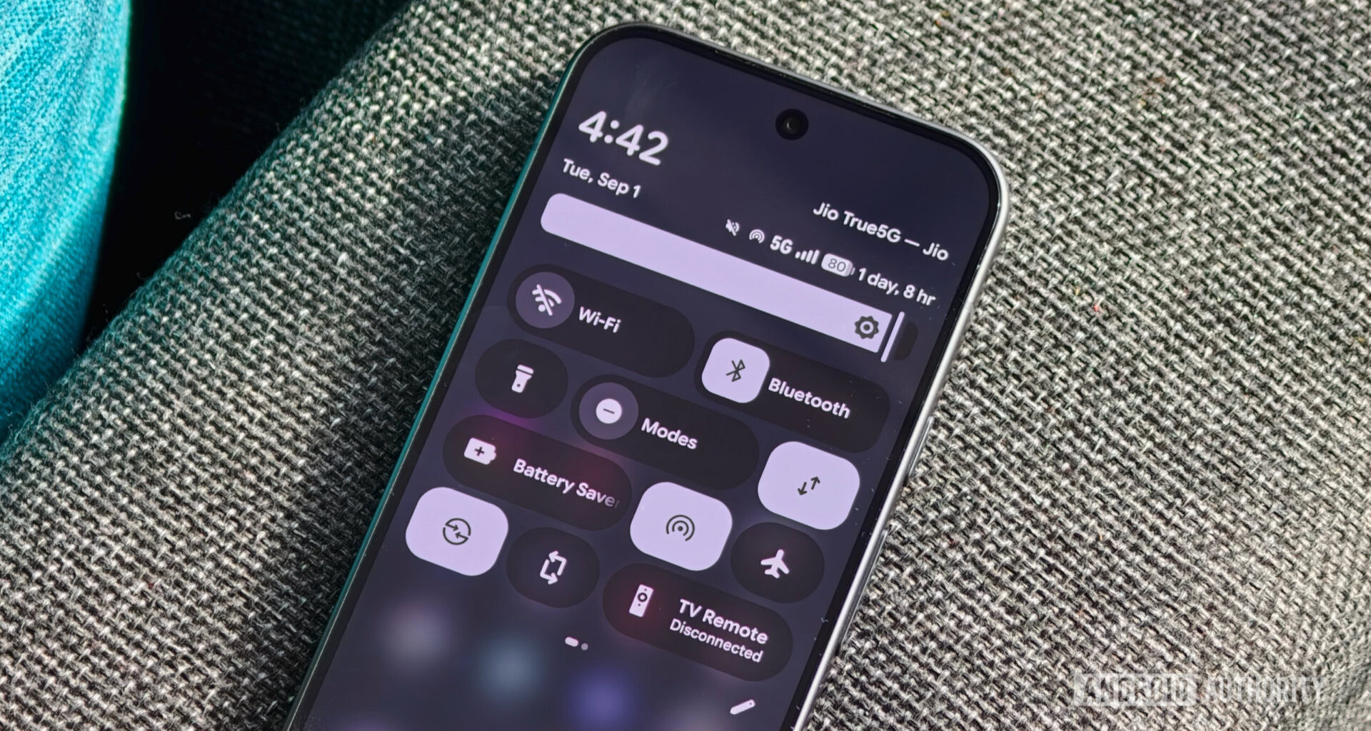 An Android smartphone resting on a textured fabric surface displaying the expanded Quick Settings shade with tiles for Wi-Fi, Bluetooth, Modes, and Battery Saver