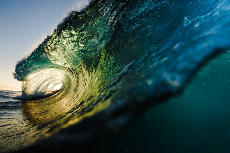 A large, translucent ocean wave curls into a barrel, illuminated by golden sunlight shining through the water.