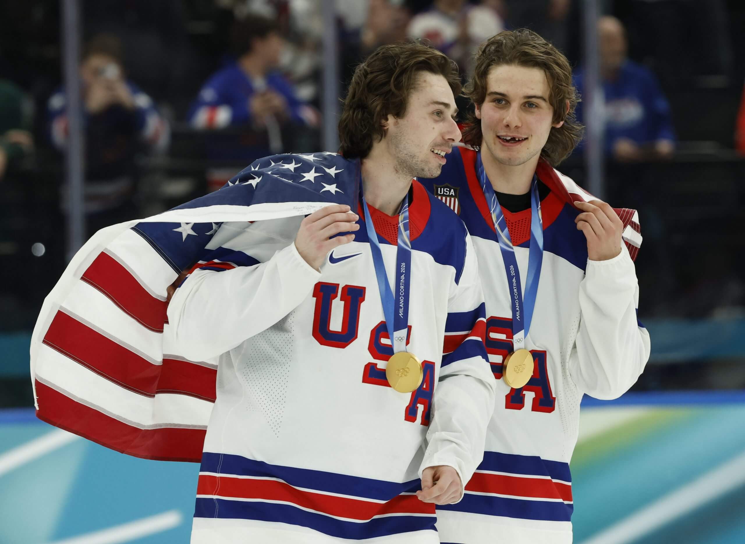 Quinn Hughes and Jack Hughes hold the U.S. flag on themselves like a cape after medaling at the Milan Cortina 2026 Olympics.