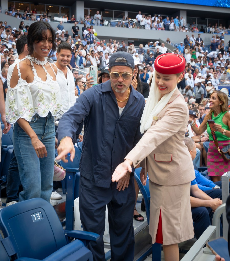 Brad Pitt and Ines de Ramon pointing at empty seats at a sporting event, with an attendant in a red hat beside them.