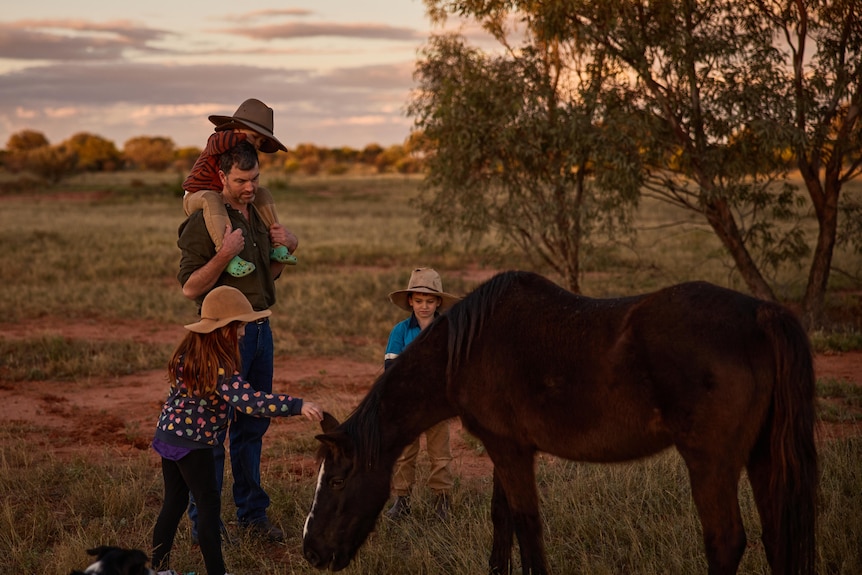 A man with his kids playing with a black horse at sunset.