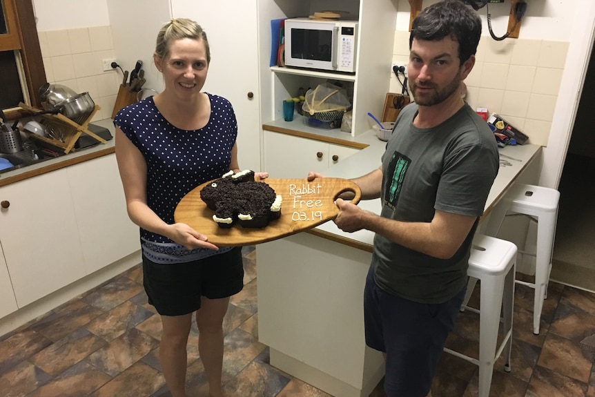 Bec West and Reece Pedler hold a cake shaped like a brown and white rabbit with words in white icing next to it: rabbit free.
