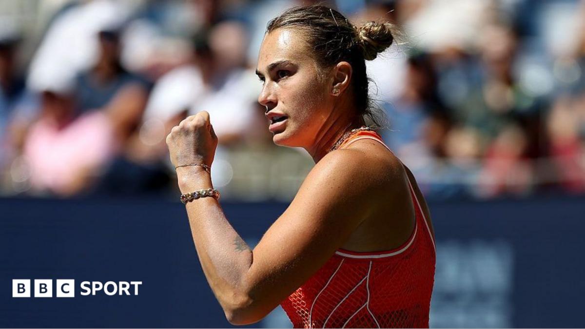 Aryna Sabalenka, wearing an orange sleeveless dress, stands side on and raises a clenched left fist