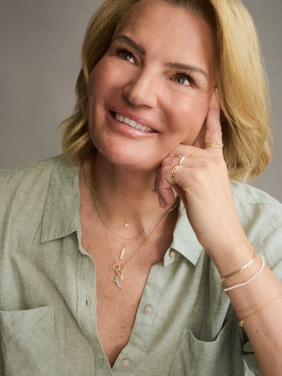 Smiling woman with layered necklaces and a relaxed green shirt, resting her chin on her hand while looking thoughtfully. Smiling woman with layered necklaces and a relaxed green shirt, resting her chin on her hand while looking thoughtfully.