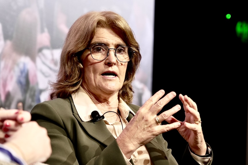 Michele Bullock, wearing glasses, a green blazer and beige shirt, speaks as she gestures with her hands.