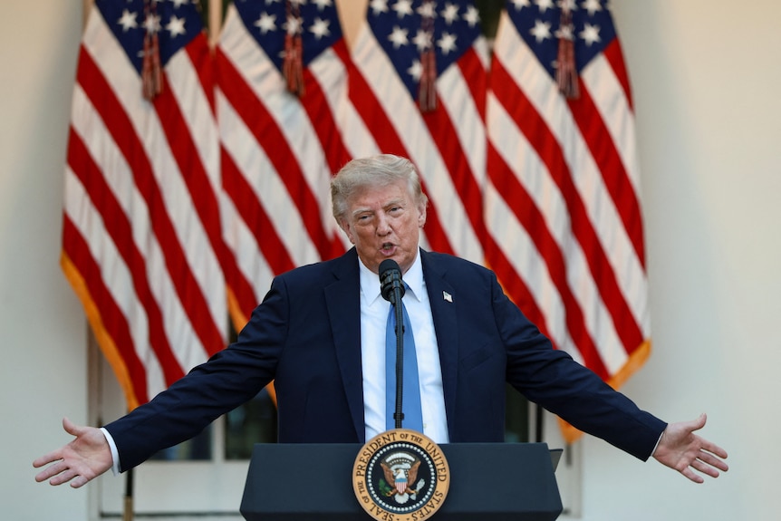 Donald Trump in a black suit, white shirt and blue tie speaking at a lectern with his arms outstretched in front of US flags.