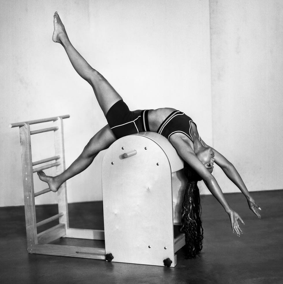 A dancer performing an expressive stretch on a Pilates reformer in a minimalistic studio setting.