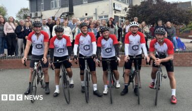 Six men in matching red and white cycling outfits sit on their bikes with the Aston Fields Social Club behind them - a two storey building with a cream base and a grey second floor. A crowd of supporters also stand behind the men.