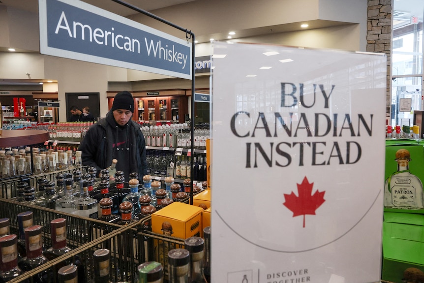 A customer in a liquor store holding a bottle. In the foreground is a sign that says 'Buy canadian instead'.