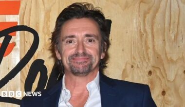 Richard Hammond is photographed from the shoulders up, standing in front of a promotional sign for the TV series 'The Grand Tour'. His face is facing slightly to the side and he is not looking at the camera. He is wearing a black suit jacket and a white shirt, with the top button undone.