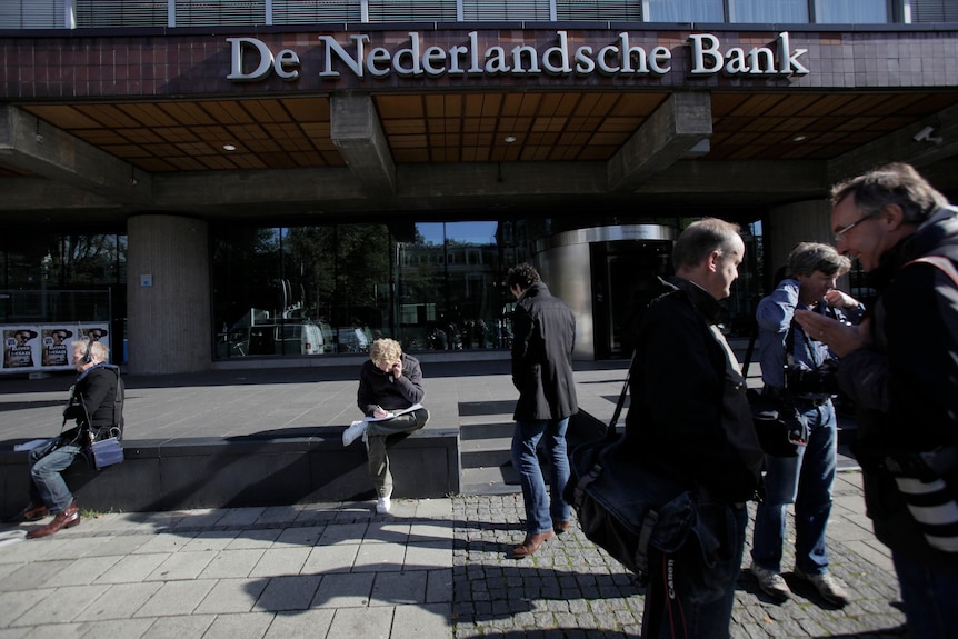 Several male reporters standing, sitting or using mobile phones outside the De Nederlandsche Bank building.