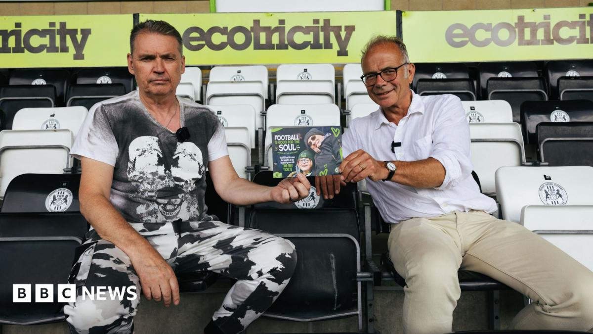 Dale Vince and Dr Simon Opher sit in a stand at Forest Green Rovers' stadium holding a flyer advertising the scheme. Vince is wearing a black and white T-shirt and black and white trousers and he looks quite serious. Opher is wearing a white shirt with the sleeves rolled up and beige trousers and he is smiling.