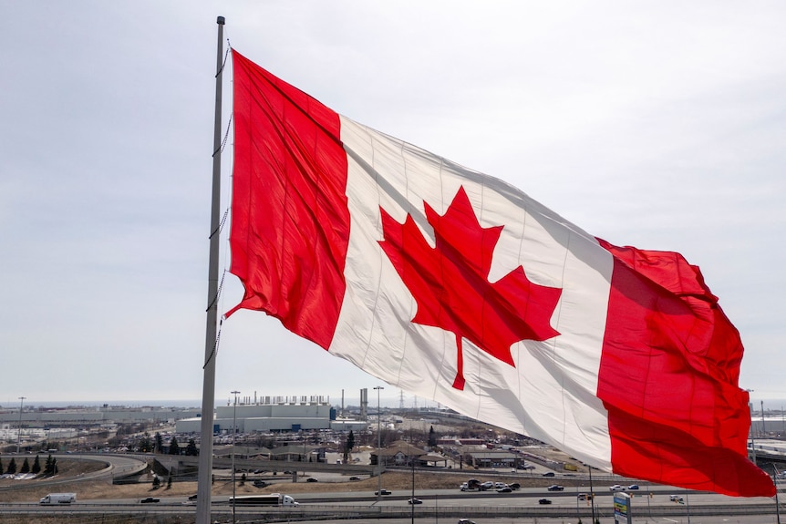 Large Canadian flag waving over a cityscape with highways and industrial buildings in the background.