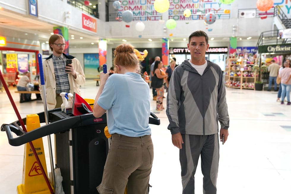 hollyoaks ste hay mall scene with people, cleaning cart, and wet floor sign.
