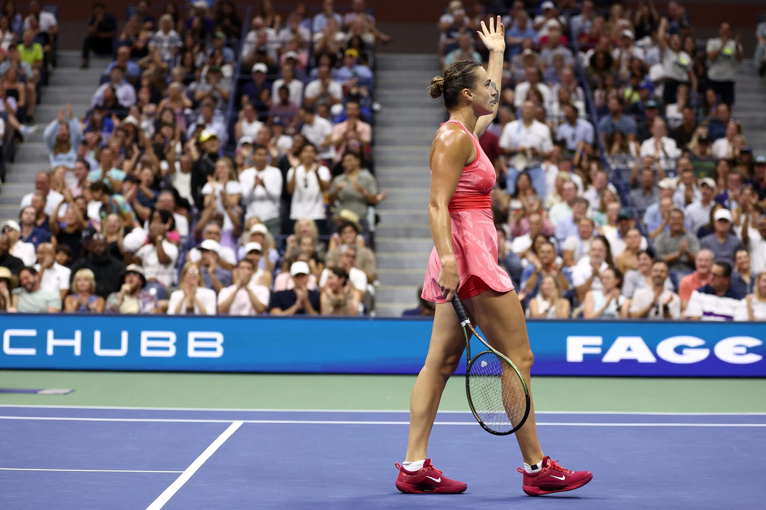 Aryna Sabalenka raises her left hand to the sky as she walks along a blue tennis court with a crowd in the background.