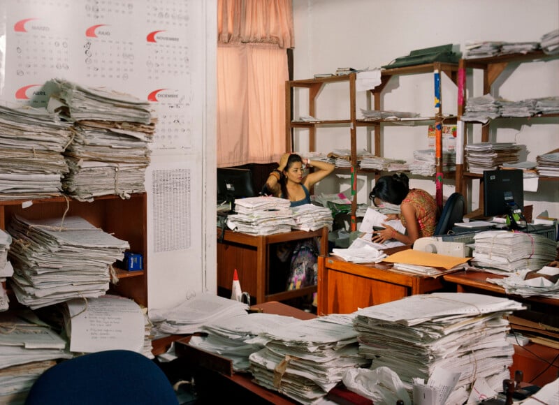 Two women work at desks in an office cluttered with large, stacked piles of paper documents and shelves filled with files.