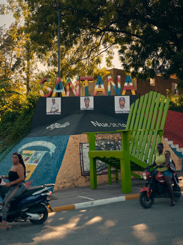 A colorful “Santana” town monument featuring photos of local baseball players and an oversize green chair, with people riding motorbikes in the foreground.