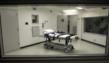 A view through the witness window into the execution chamber, showing a gurney with leather restraint straps and an adjacent room with folding chairs.