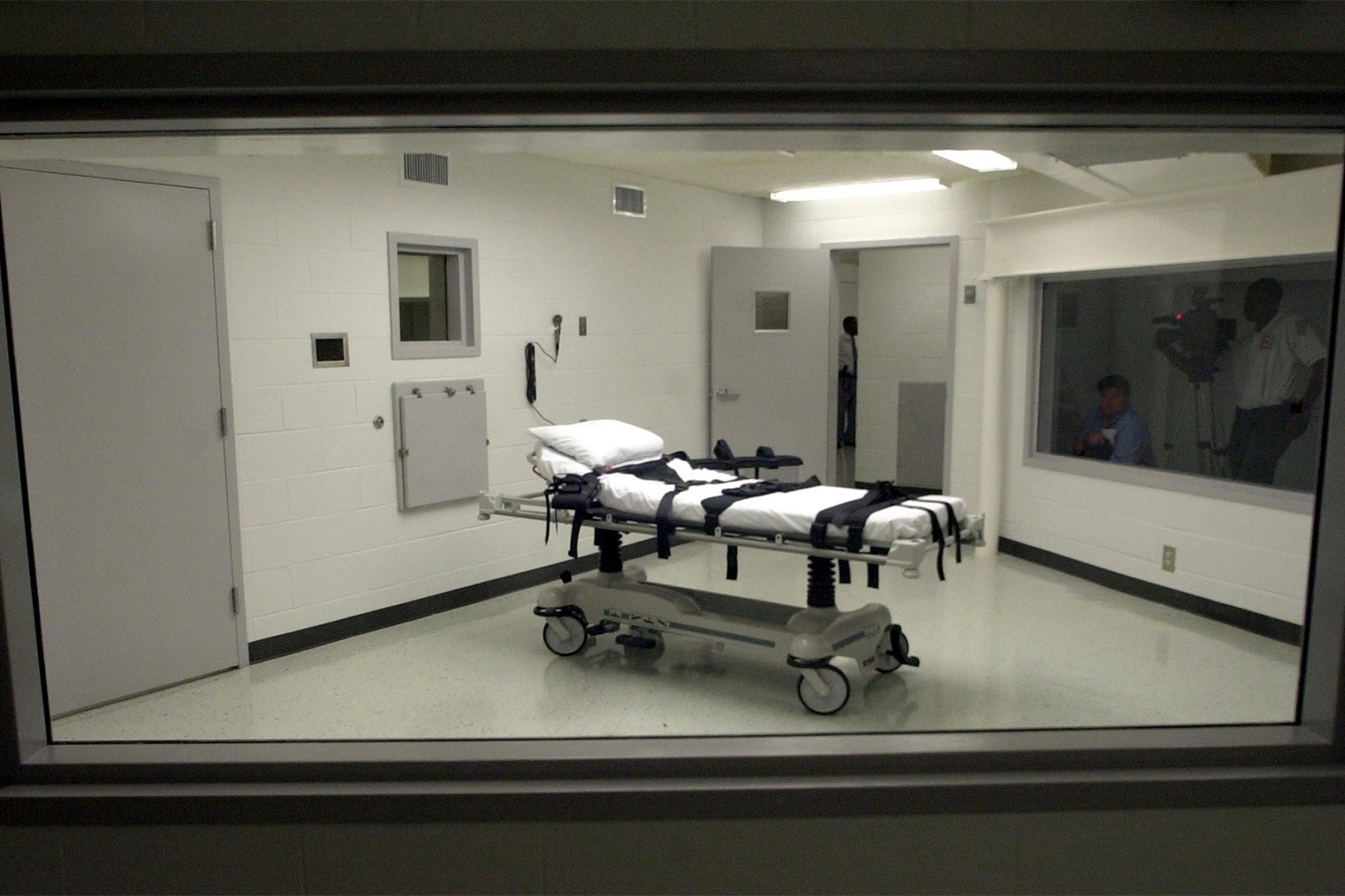 A view through the witness window into the execution chamber, showing a gurney with leather restraint straps and an adjacent room with folding chairs.