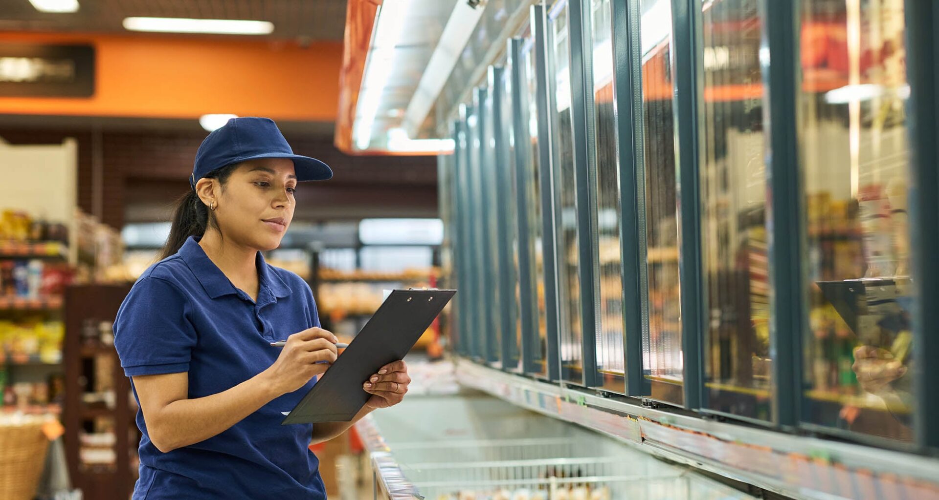 A middle aged woman working at a grocery store stands in front of a freezer with a clipboard.