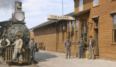 A steam locomotive numbered 245 is stopped at a wooden train station labeled Tomahawk, with six men standing nearby on the platform.
