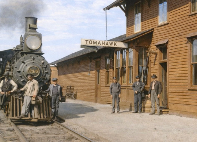 A steam locomotive numbered 245 is stopped at a wooden train station labeled Tomahawk, with six men standing nearby on the platform.
