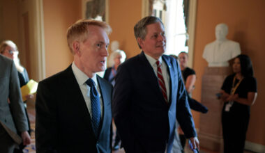 Sen. James Lankford and Sen. Steve Daines in suits walk through a hallway with others in the background. A white statue is visible on a pedestal, and sunlight streams through windows, casting a warm glow on the scene.