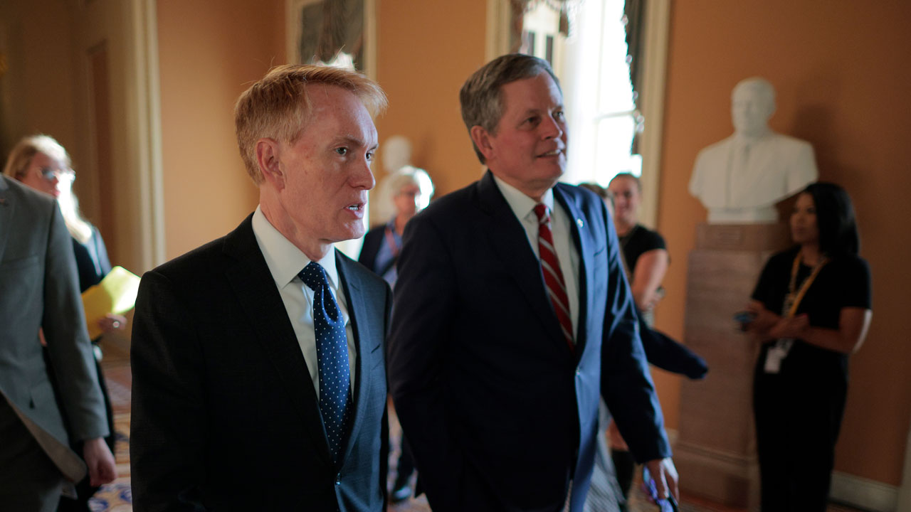 Sen. James Lankford and Sen. Steve Daines in suits walk through a hallway with others in the background. A white statue is visible on a pedestal, and sunlight streams through windows, casting a warm glow on the scene.