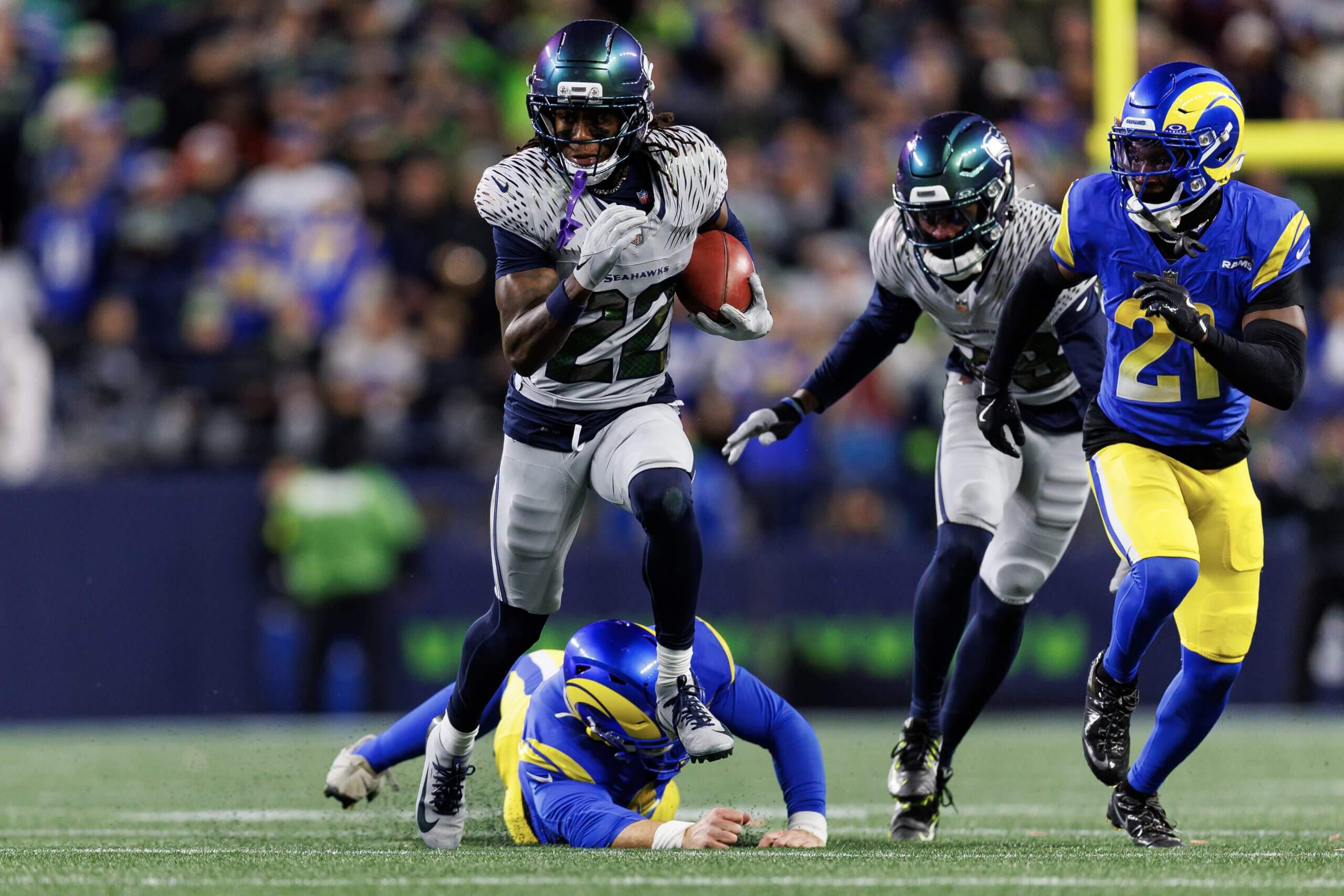 Rashid Shaheed, wearing a gray No. 22 Seahawks uniform, tucks the ball under his left arm while running away from two Rams defender, including one who is sprawled on the ground.