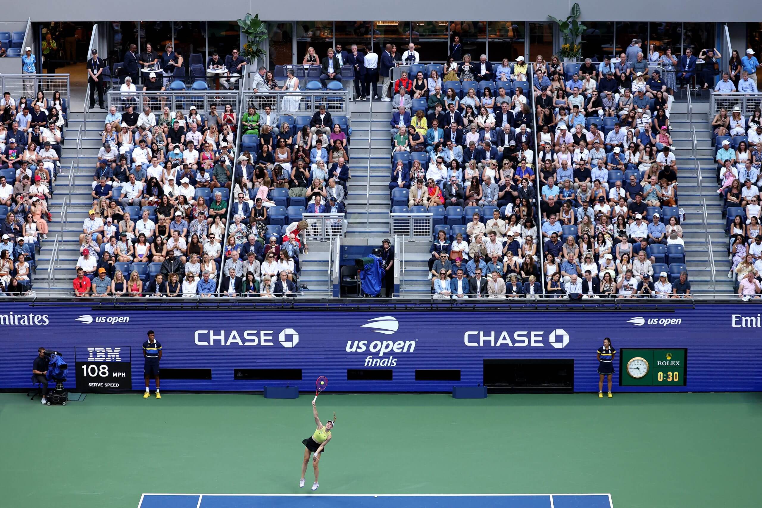 Elena Rybakina serves in front of a tennis stadium.
