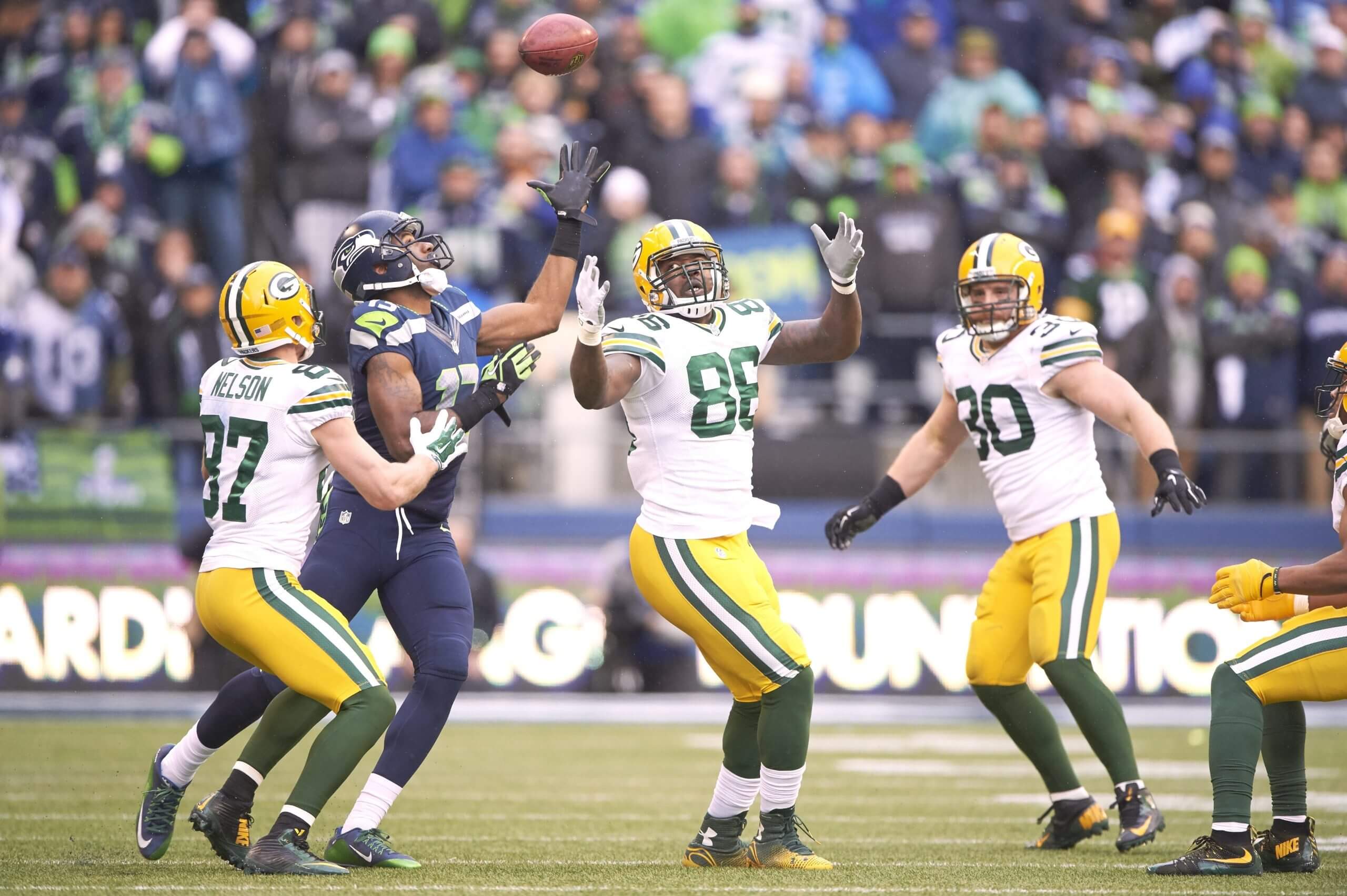 Brandon Bostick, wearing a white No. 86 Packers uniform, holds both hands up while looking at the ball in the air as Seahawks receiver Chris Matthews, wearing a navy No. 13 uniform, reaches to catch it in front of Jordy Nelson.