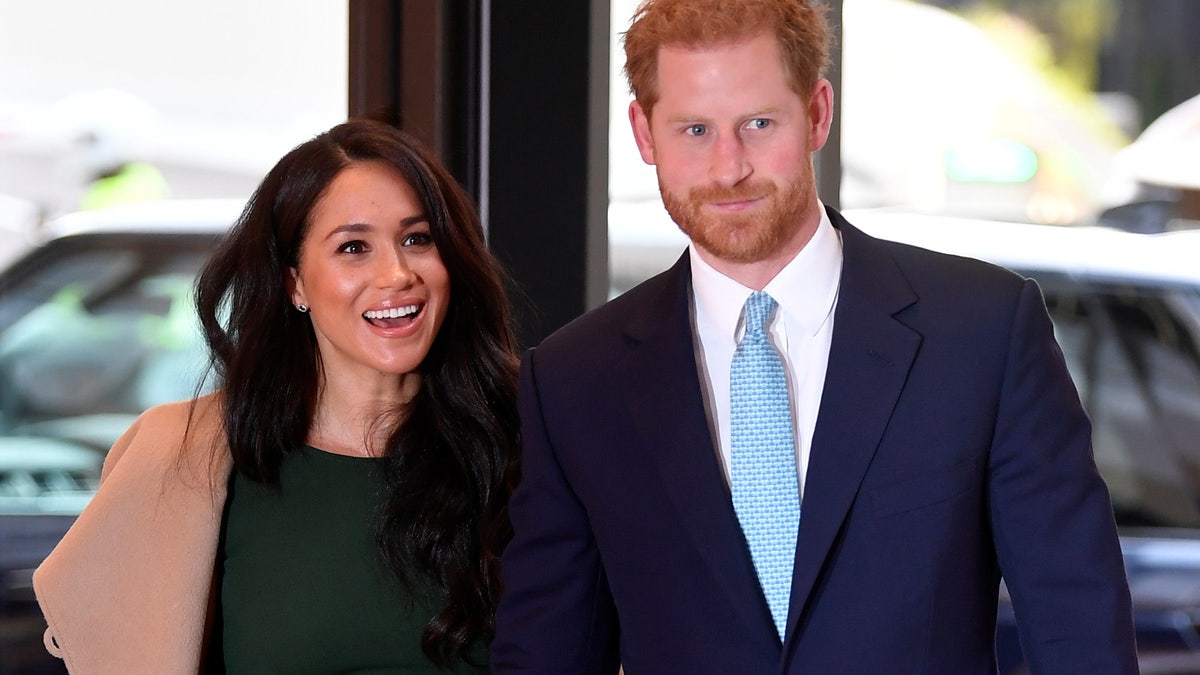 Meghan Markle in a green dress and camel colored jacket walks with Prince harry in a navy suit and teal tie