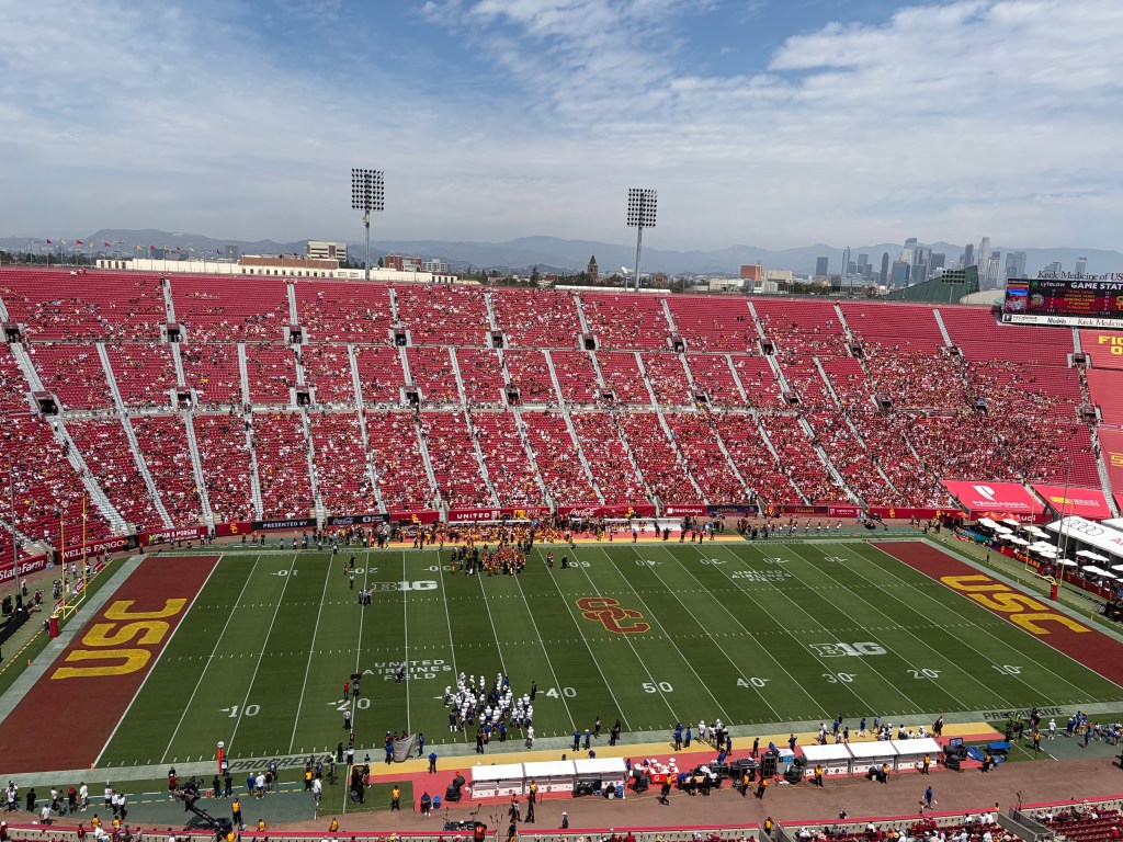 An overhead view of a football stadium during a game, with red seats filled with spectators, players on the field, and a city skyline in the background.