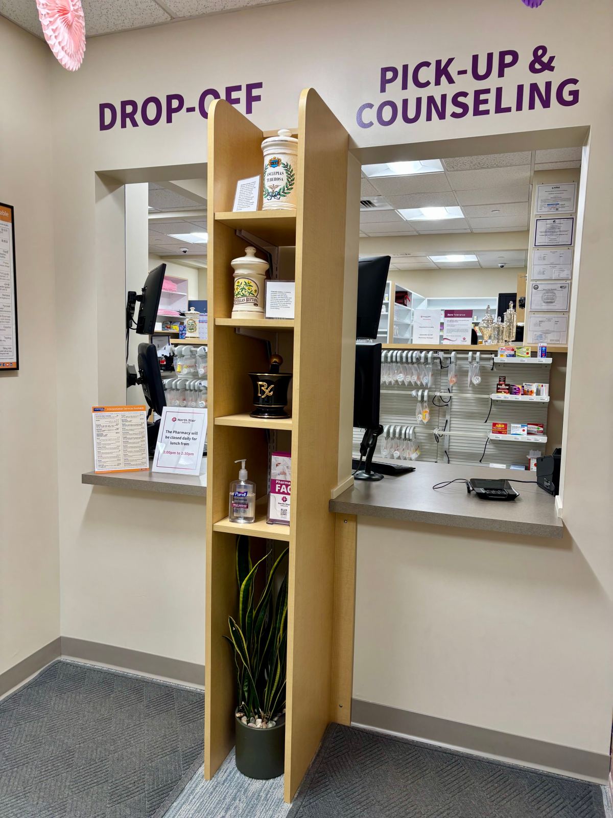 Pharmacy counter with separate drop-off and pick-up windows, separated by a wooden shelf holding decorative jars, a plant, and hand sanitizer bottles.