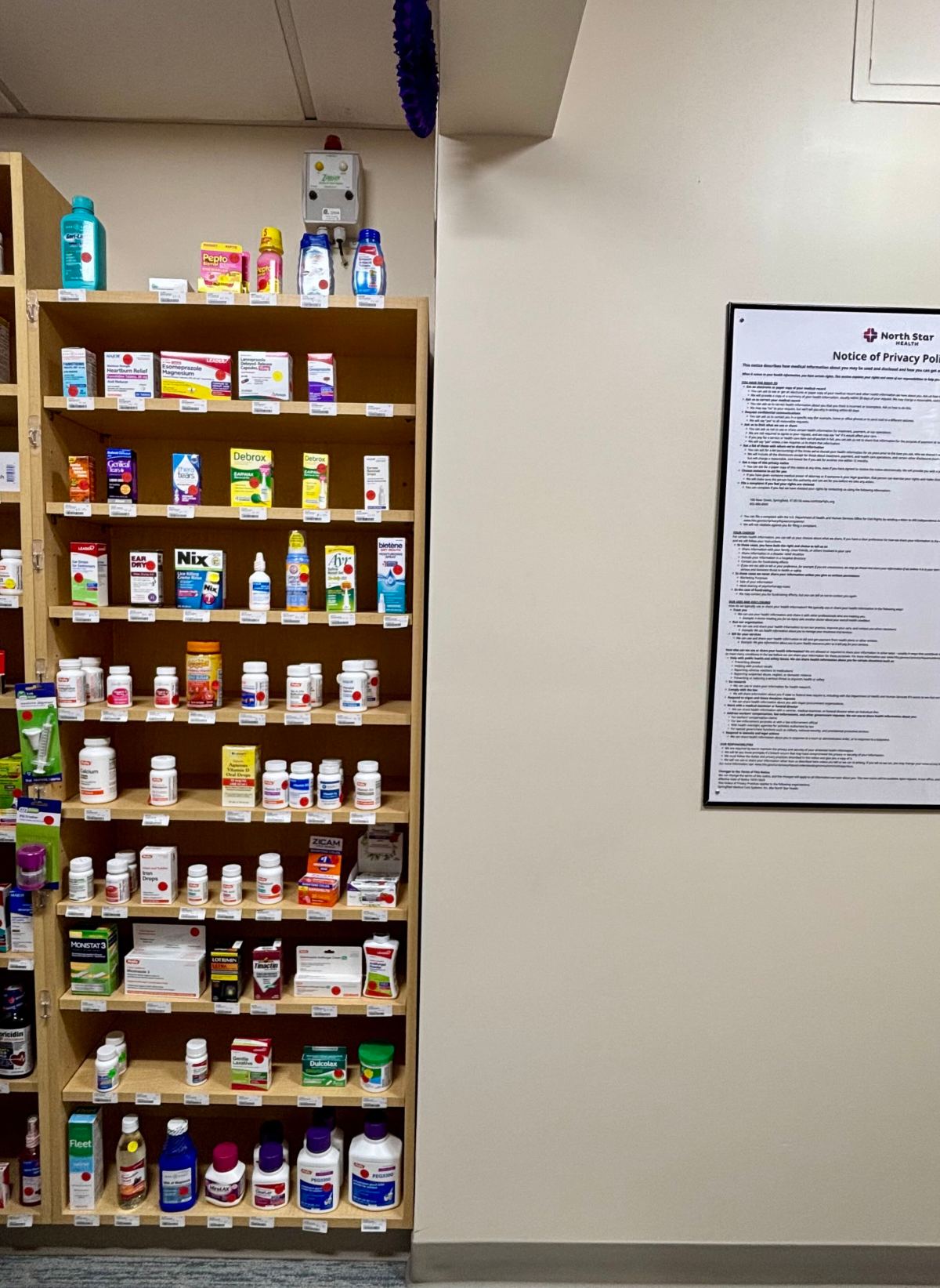 Wooden shelves filled with various medications and health products next to a beige wall with a framed “Notice of Privacy Practices” document.