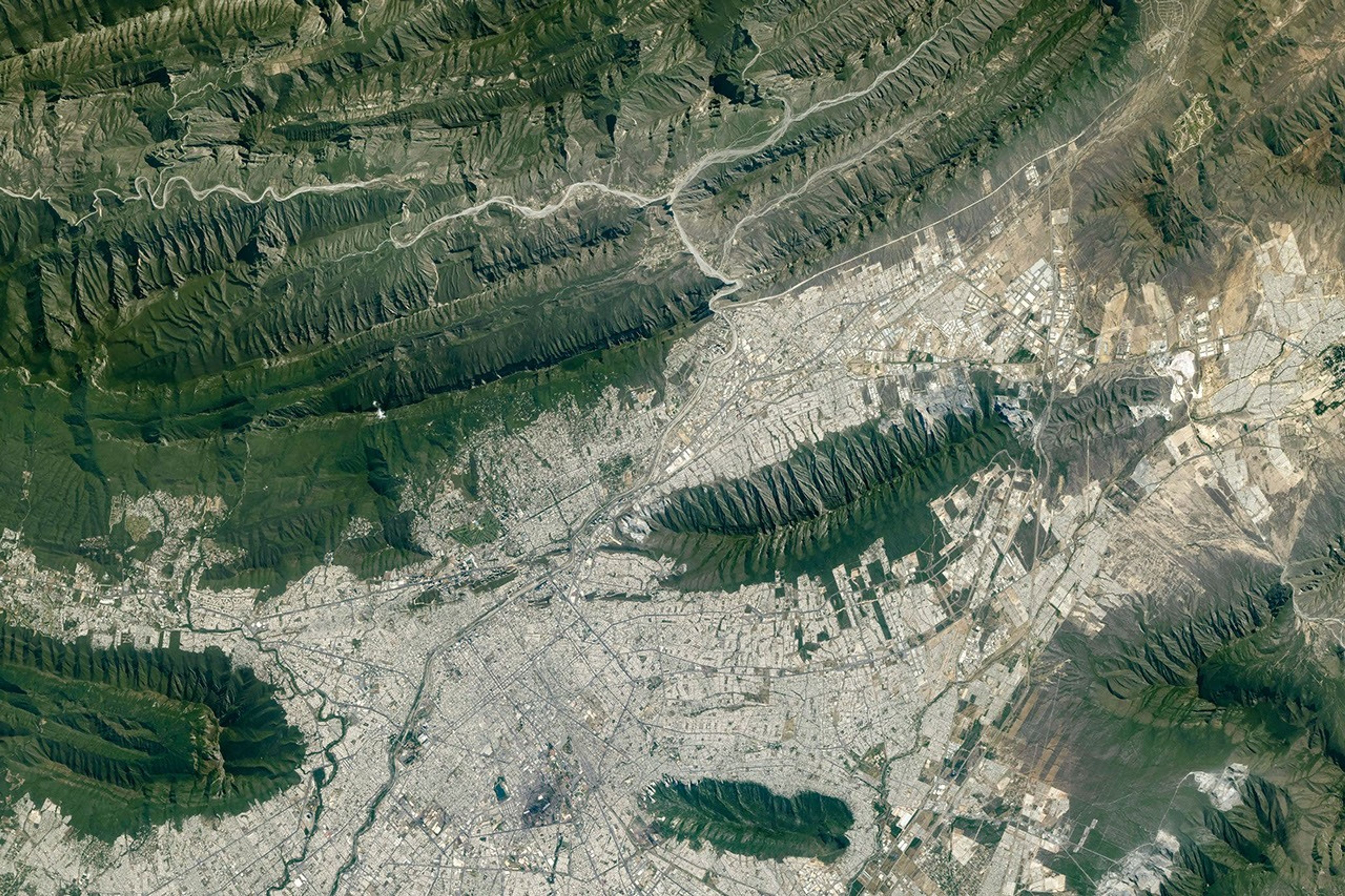 The light-colored urban development of Monterrey, Mexico, fills most of the top half of the photo, and green parallel mountain ridges arc across the bottom of the frame.