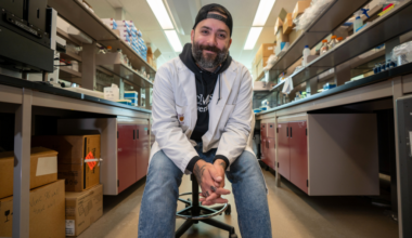Jon Stokes, wearing a ball cap, a lab coat, a hoodie and jeans, sits in the middle of his lab, smiling down at the camera.