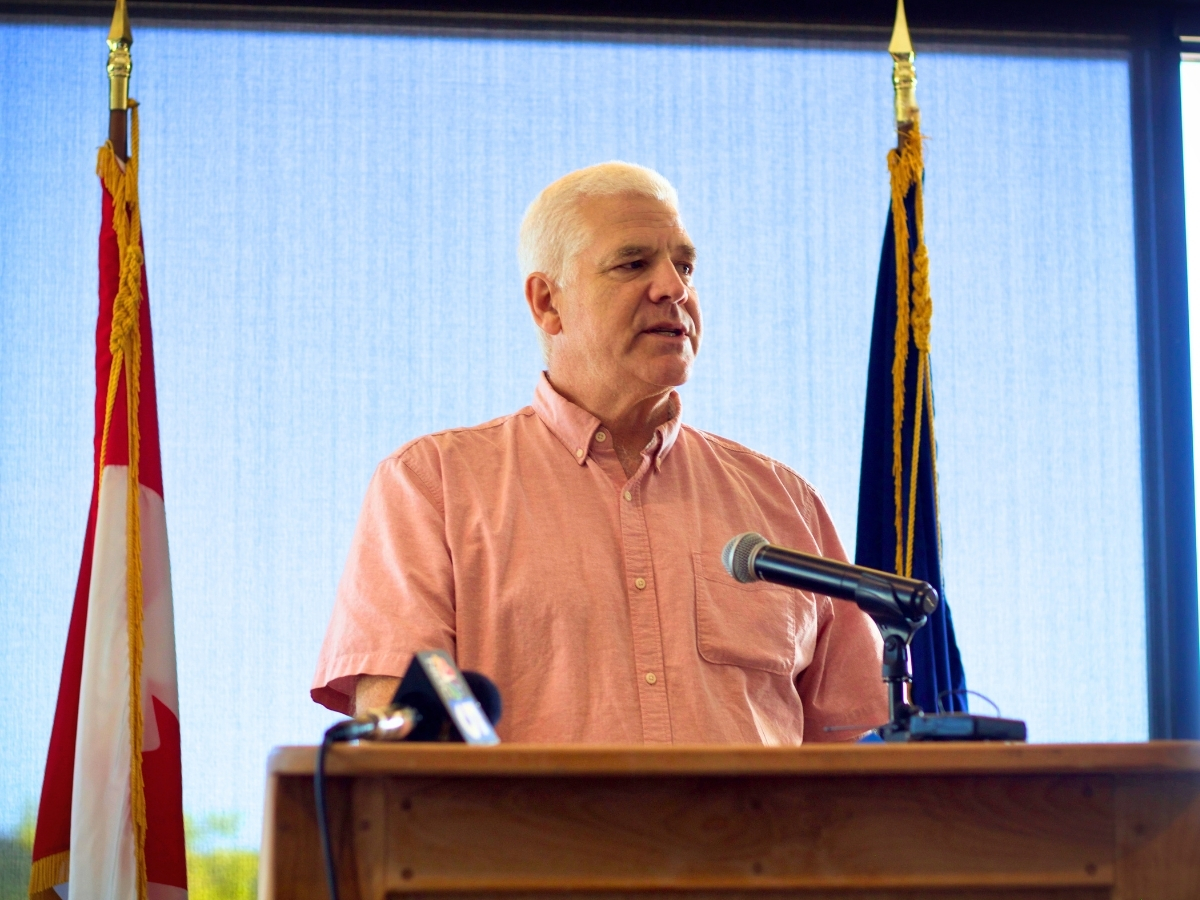 A man in a pink shirt stands at a podium with microphones, flanked by two flags, speaking in front of a blue curtain.
