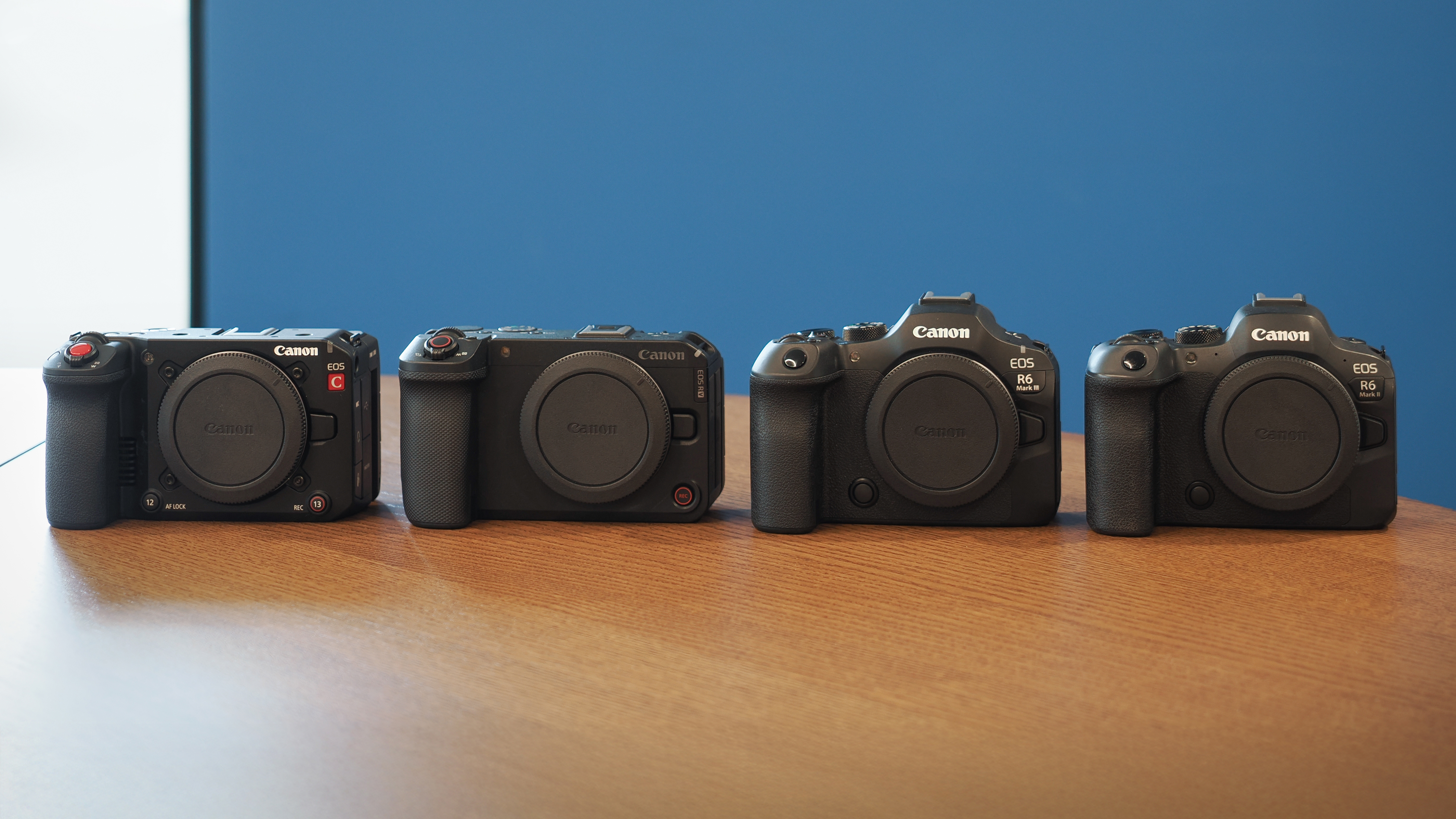 The Canon EOS C50, R6 V, R6 Mark III and R6 Mark II on a wooden surface, against a blue background