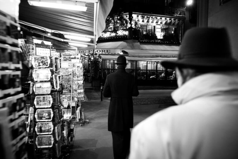 A person in a hat and coat walks toward the Café de Flore at night, viewed from behind another person in the foreground.