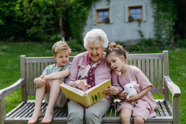 Portrait two siblings with grandmother, sitting on bench in the garden, reading book together. Grandson and granddaughter enjoying together time with elderly grandma, looking at photos from photo album.