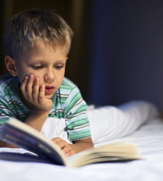 Little boy aged 6 is reading book in bed before going to sleep. The book is very interesting and the boy is quite lost in it.