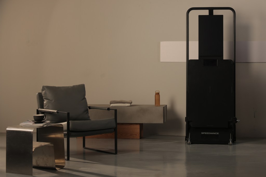 A folded black Speediance smart home gym stands upright next to an armchair.