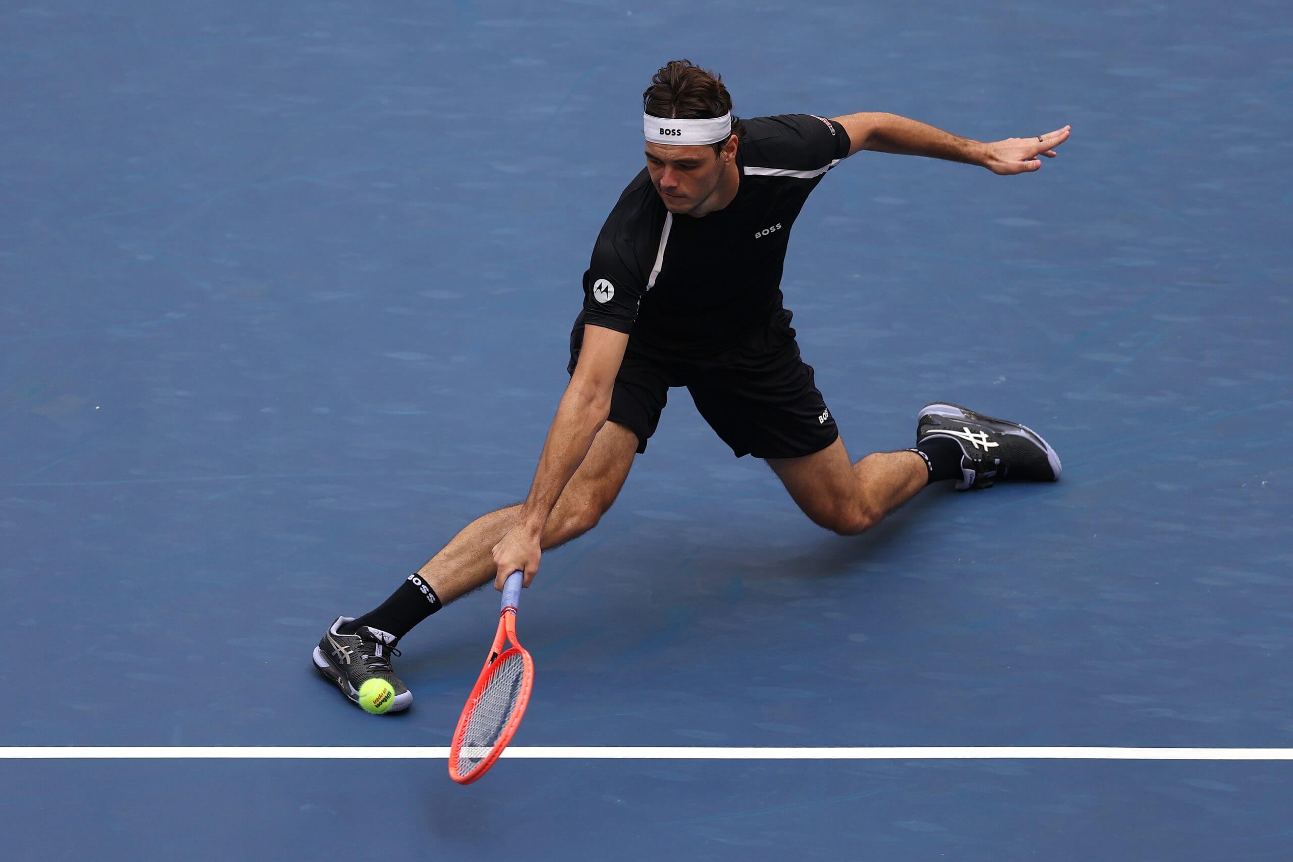 Taylor Fritz stretches for a a short backhand on a blue tennis court.