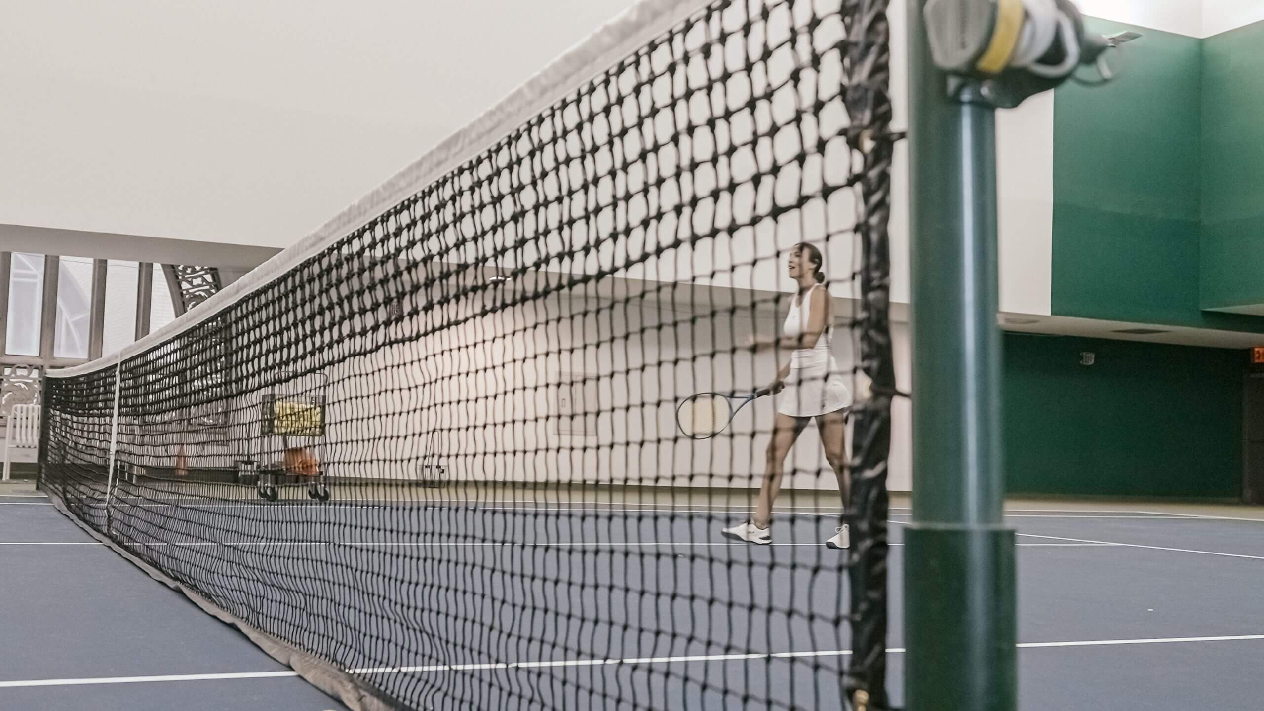 A woman stands on a tennis court with an ornate arched window across from its net.