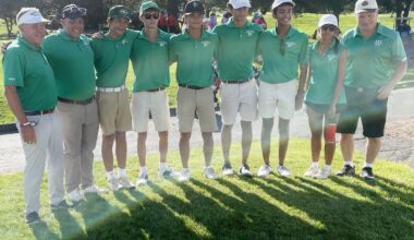 Group of golfers in green polo shirts posing together on a sunny course, arms around each other.