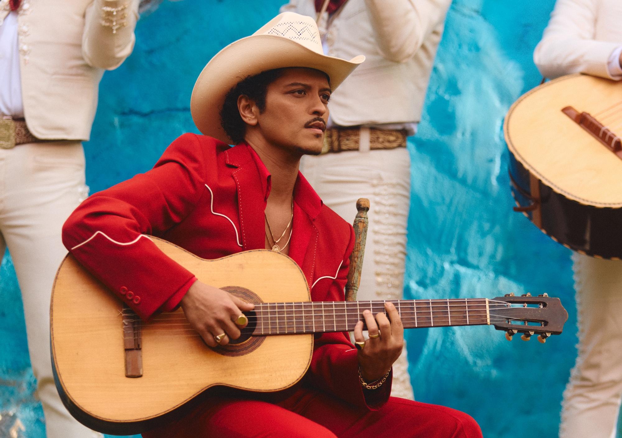 Photo of Bruno Mars wearing a red suit, a white hat, and holding a guitar.