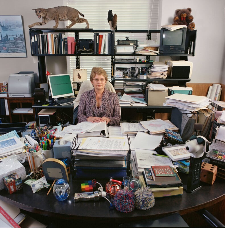 A woman sits at a cluttered desk in an office, surrounded by stacks of papers, office supplies, a computer, and various decorative items.