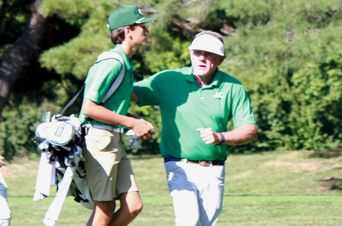 Two golfers on a sunny course shake hands; a young player with a golf bag walks beside an older coach in a visor.