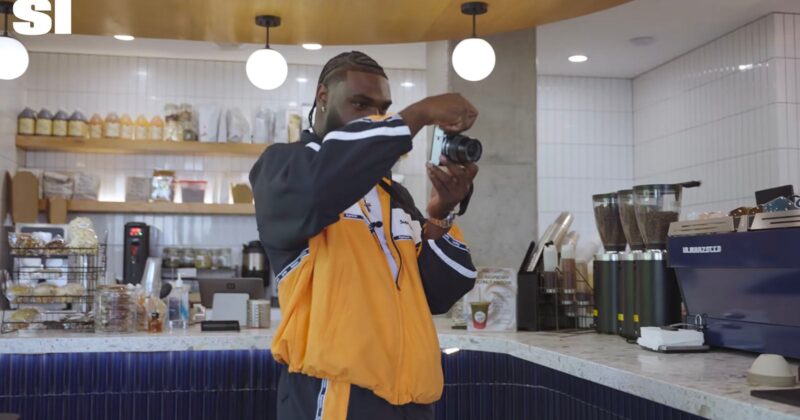 A man with braided hair wearing a black and orange jacket holds a camera up to his face to take a picture in a cafe.